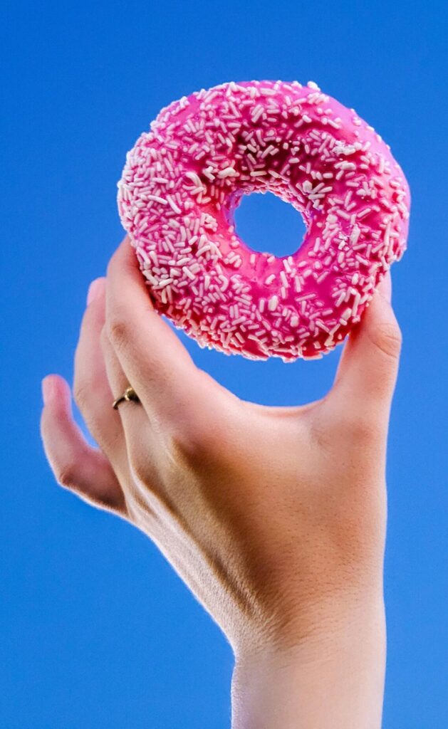 A hand holds a pink frosted donut with white sprinkles against a vibrant clear blue sky, offering a playful and colorful composition.